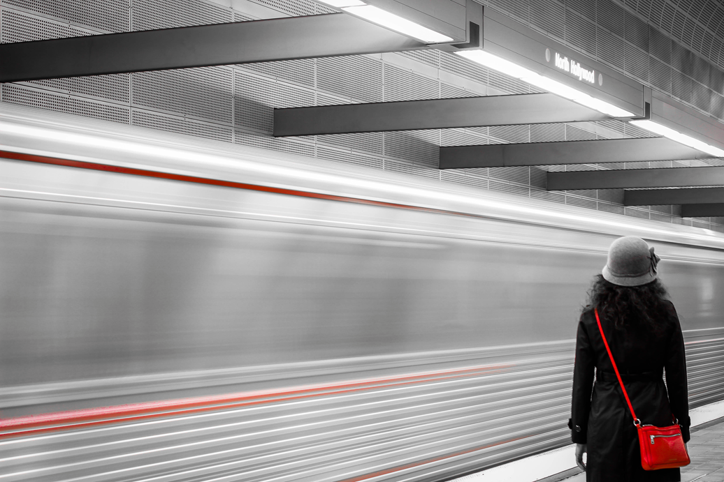 Woman in grey hat and black trench coat with red purse standing as training moving behind her.